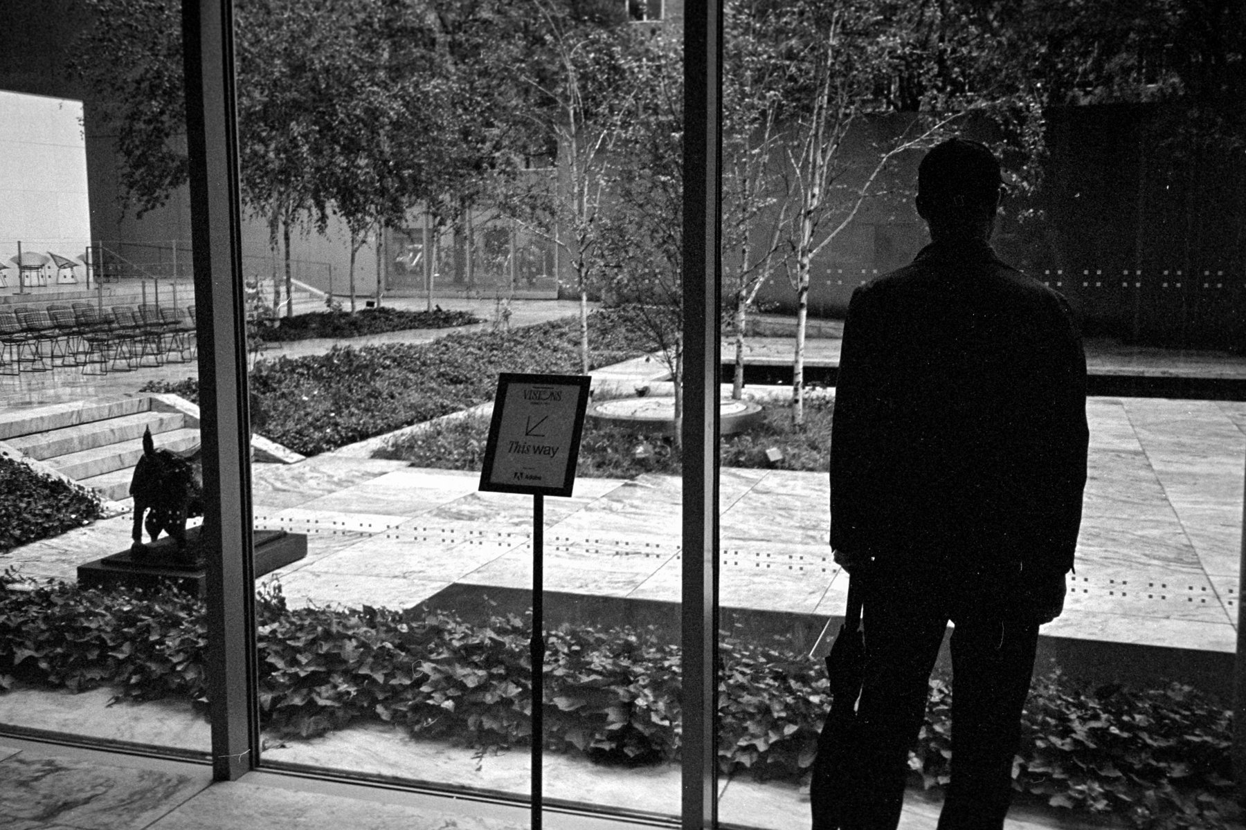 A guest looks through a MoMA window toward the sculpture garden, beside a VISIONS sign